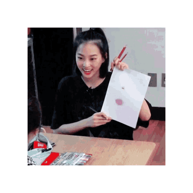a girl is sitting at a table holding a piece of paper and two pencils