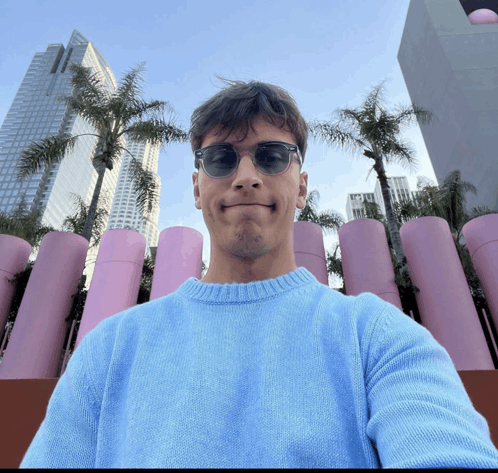 a man wearing sunglasses and a blue sweater takes a selfie in front of pink pipes