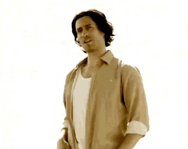a man in a tan shirt and white tank top is standing with his hands in his pockets