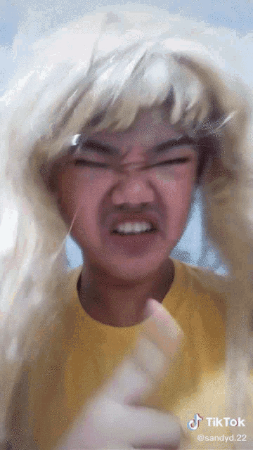 a man wearing a blonde wig and a yellow shirt is making a funny face with tiktok written below him
