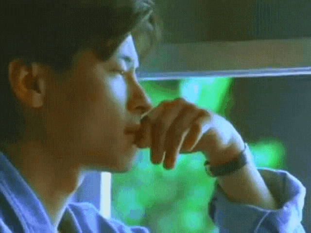 a man in a blue shirt is looking out a window with his hand on his chin .