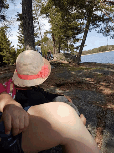 a woman wearing a hat with a flower on it sits on a rock near a lake