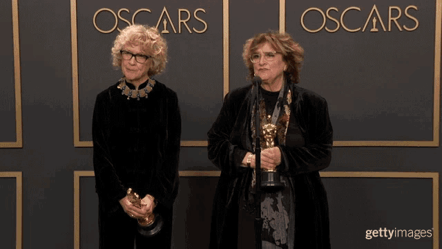 two women holding trophies in front of a wall that says oscars