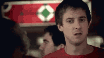 a man in a red shirt is making a funny face while standing in front of a red sign .