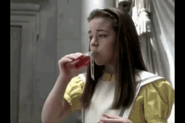 a girl in a yellow dress and white apron is drinking from a bottle .