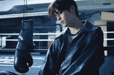 a man in a leather jacket sits in a boxing ring with a pair of boxing gloves hanging from a rope