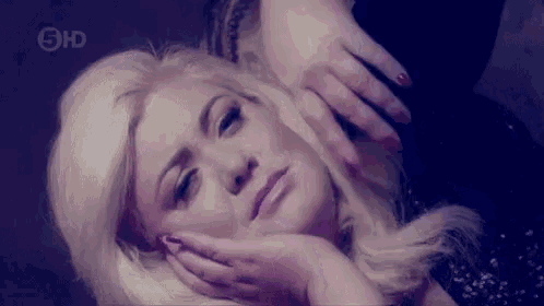 a woman with blonde hair is laying down with her hands on her face and looking at the camera .