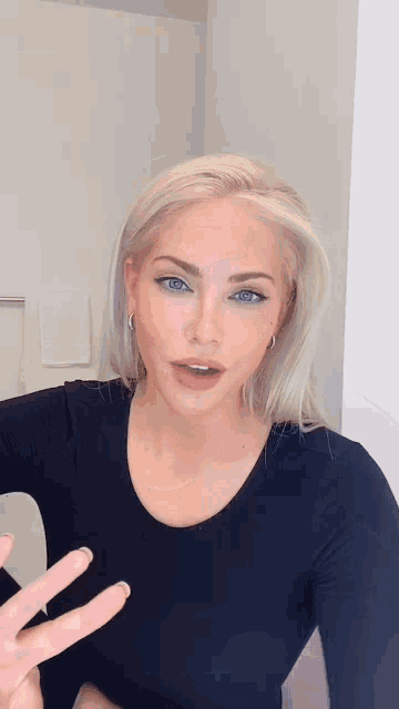 a woman with blonde hair and blue eyes is wearing a black shirt and giving the peace sign