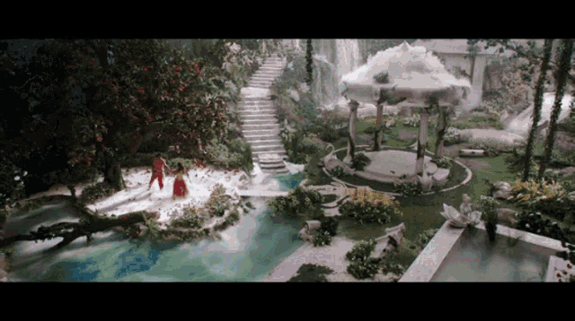 a man and a woman are walking in a garden with a waterfall in the background