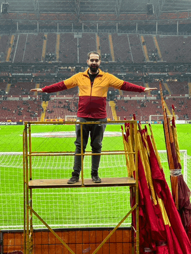 a man in an orange and red jacket stands on a scaffolding in front of a stadium that says uefa europa league
