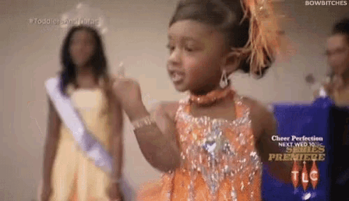 a little girl in an orange dress is giving a middle finger while standing next to a woman in a yellow dress .
