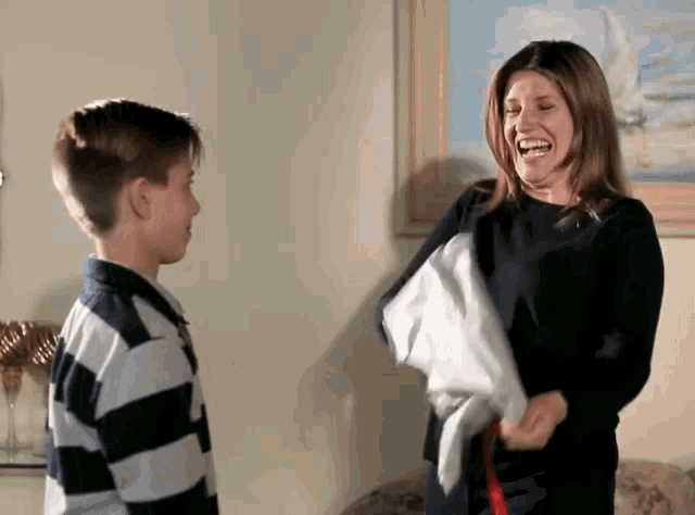 a woman laughs while holding a white shirt in front of a boy in a striped shirt