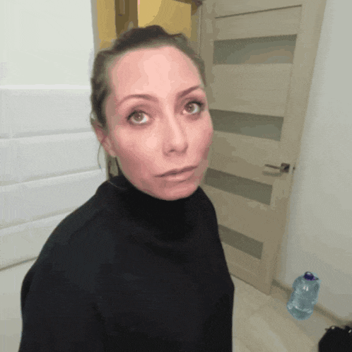 a woman in a black turtleneck looks at the camera in front of a door