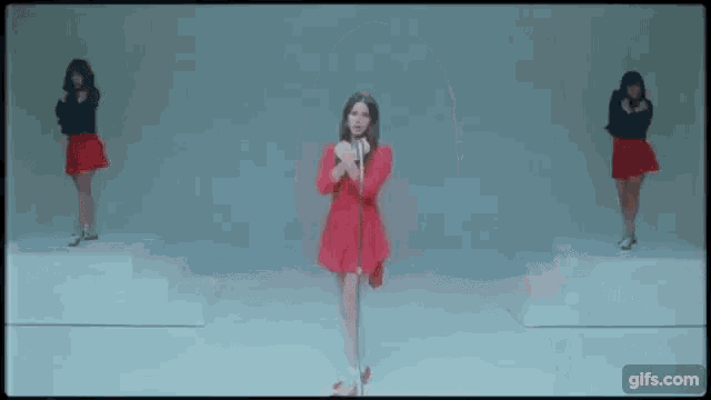 a woman in a red dress is singing into a microphone while three women in red skirts are dancing in the background .