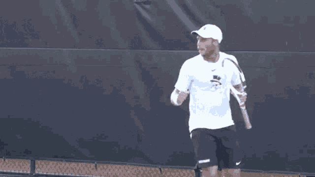 a man wearing a white hat and a white shirt is pointing at something while standing on a tennis court .