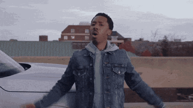 a man in a denim jacket is standing in front of a white car with his arms outstretched