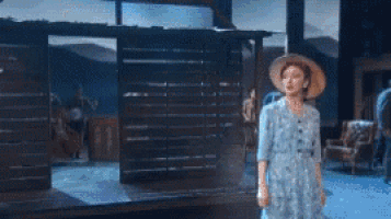 a woman in a blue dress and straw hat is dancing in front of a wooden building .