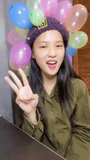a girl wearing a purple hat with a crown on it is surrounded by balloons and making a peace sign
