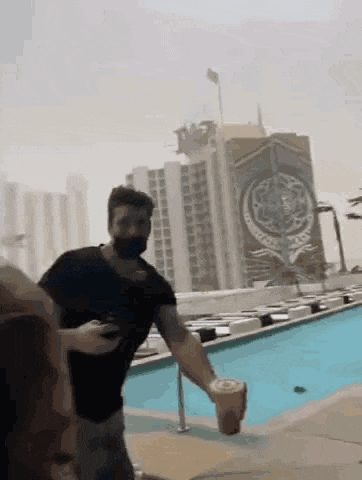 a man wearing a mask is standing next to a pool holding a cup of coffee .