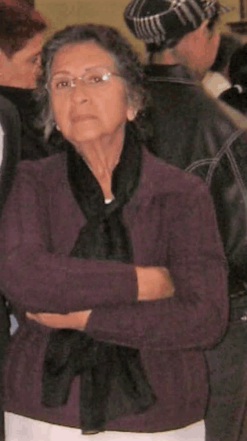 a woman wearing a purple jacket and a black scarf stands with her arms crossed