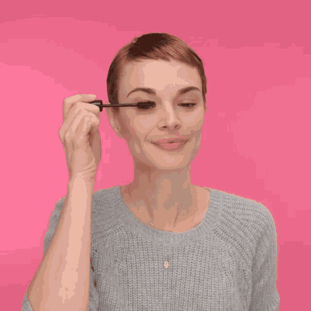 a woman wearing a grey sweater is applying mascara