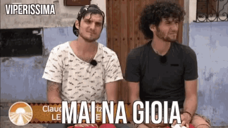two men are sitting next to each other with the words mai na gioia on the bottom right