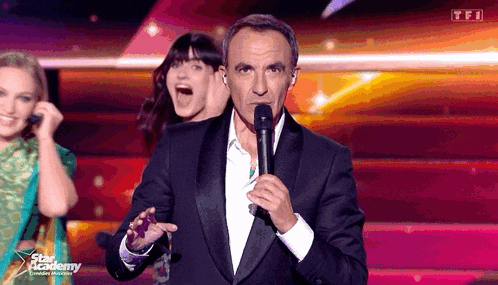 a man in a tuxedo is holding a microphone in front of a tfi logo on the screen