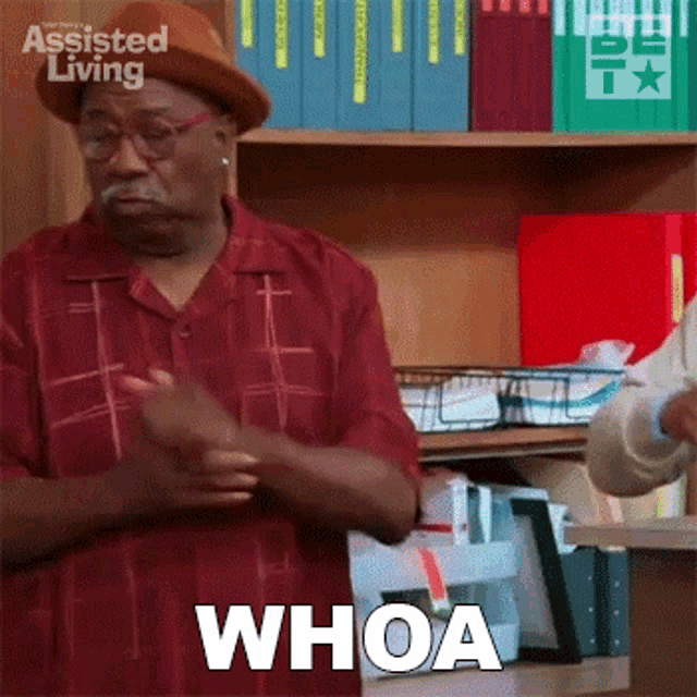 a man wearing a hat and glasses is standing in front of a bookshelf and making a funny face .