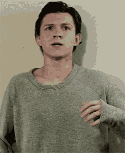 a young man in a grey sweater is standing in front of a wall and making a funny face .