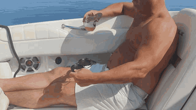 a shirtless man is sitting on a boat holding a glass