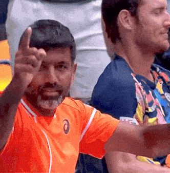 a man with a beard wearing an orange asics shirt is giving the middle finger