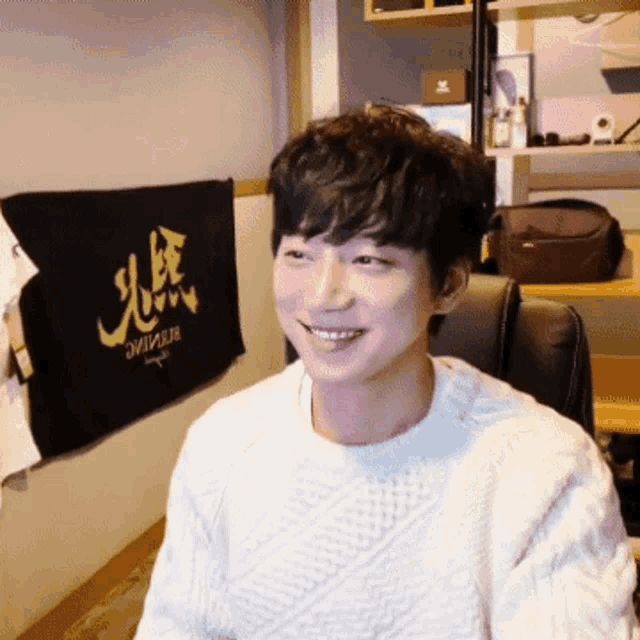 a man wearing a white sweater is smiling in front of a black towel that says ' ae ' on it