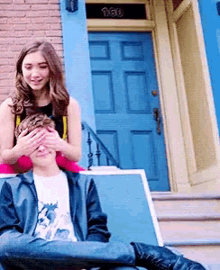 a girl covering a boy 's eyes with her hands in front of a blue door