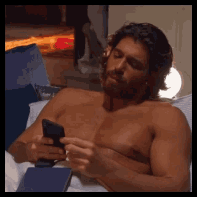a shirtless man is laying on a bed using a cell phone