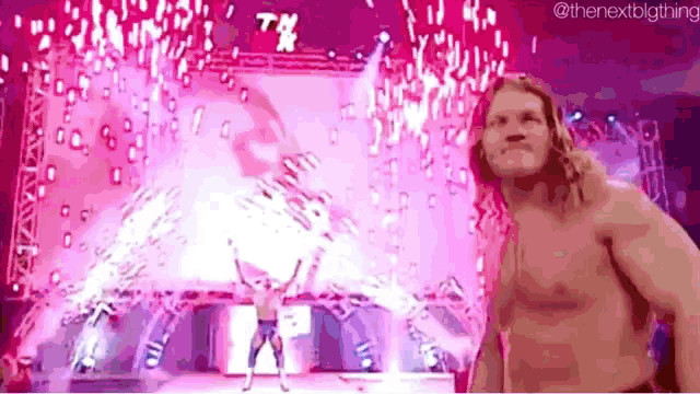 a man without a shirt is standing on a stage in front of a pink background with fireworks .