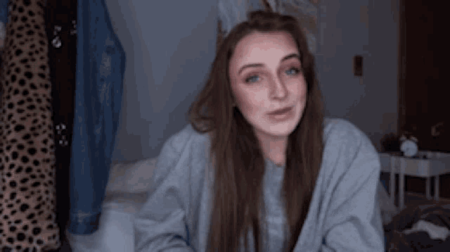 a woman is sitting on a bed wearing a grey sweater and smiling .