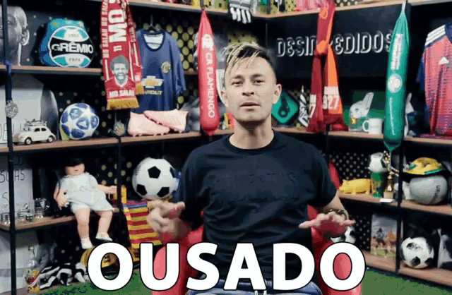 a man is sitting in front of a shelf full of soccer balls and says " ousado "