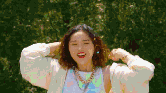 a woman wearing a white shirt and a necklace smiles with her hands in her hair