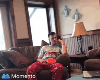a man in pajamas is sitting on a couch with a momento icon in the corner