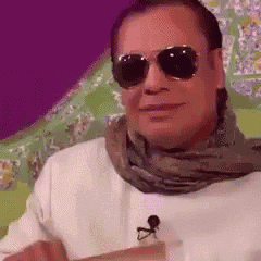 a man wearing sunglasses and a scarf is sitting in a chair and smiling .