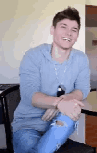 a young man in a blue shirt and ripped jeans is sitting at a table with his legs crossed and smiling .