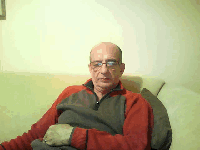 a bald man wearing glasses and a red and grey sweater is sitting on a couch