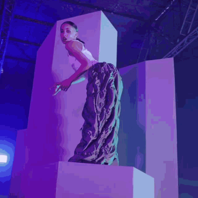 a woman in a white top and camo pants stands on a purple cube