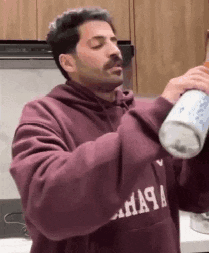 a man in a maroon hoodie is pouring a drink from a can into his mouth .