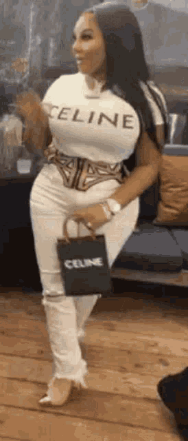 a woman wearing a celine t-shirt and white pants is standing in a room holding a celine bag .