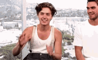 a man in a white tank top and suspenders is standing next to another man in a white shirt and smiling .