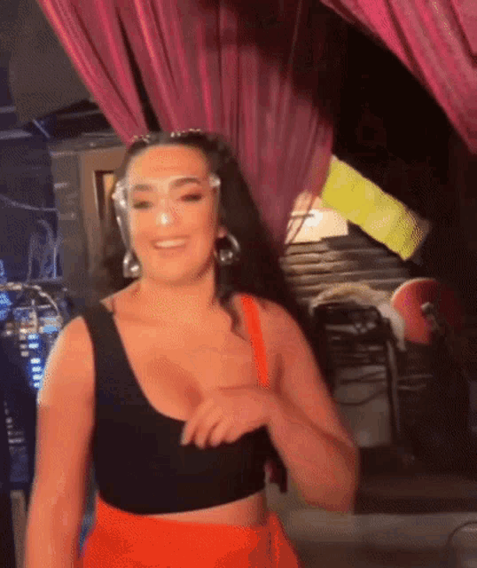 a woman wearing a black crop top and orange shorts is smiling while standing in front of a pink curtain .
