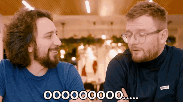 two men are sitting next to each other and one of them is wearing glasses and the other is wearing a turtleneck