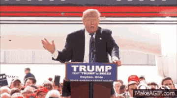 a man in a suit and tie stands at a podium holding a trump sign