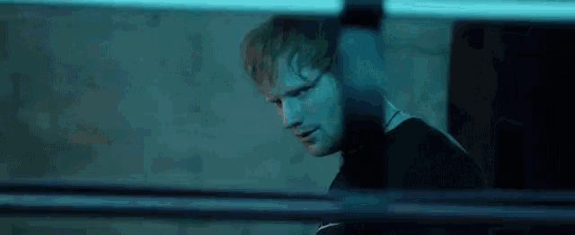 ed sheeran is standing in a dark room behind a boxing ring looking at the camera .